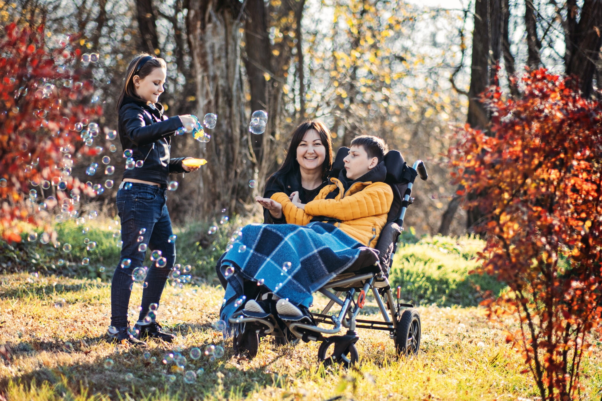 Mother and sibling playing with bubbles with child in wheelchair, inclusive outdoor play, disability support, family bonding, adapted activities.